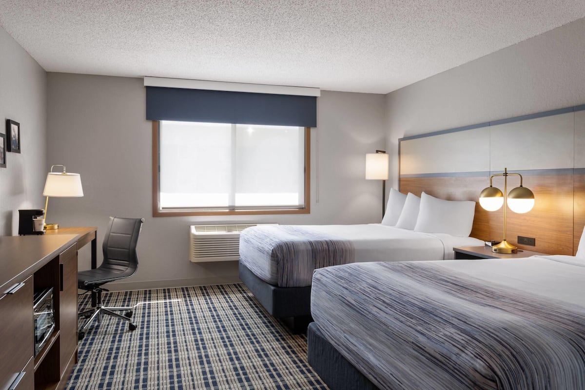 A hotel room with two beds, a desk, a chair, a lamp, and a large window.