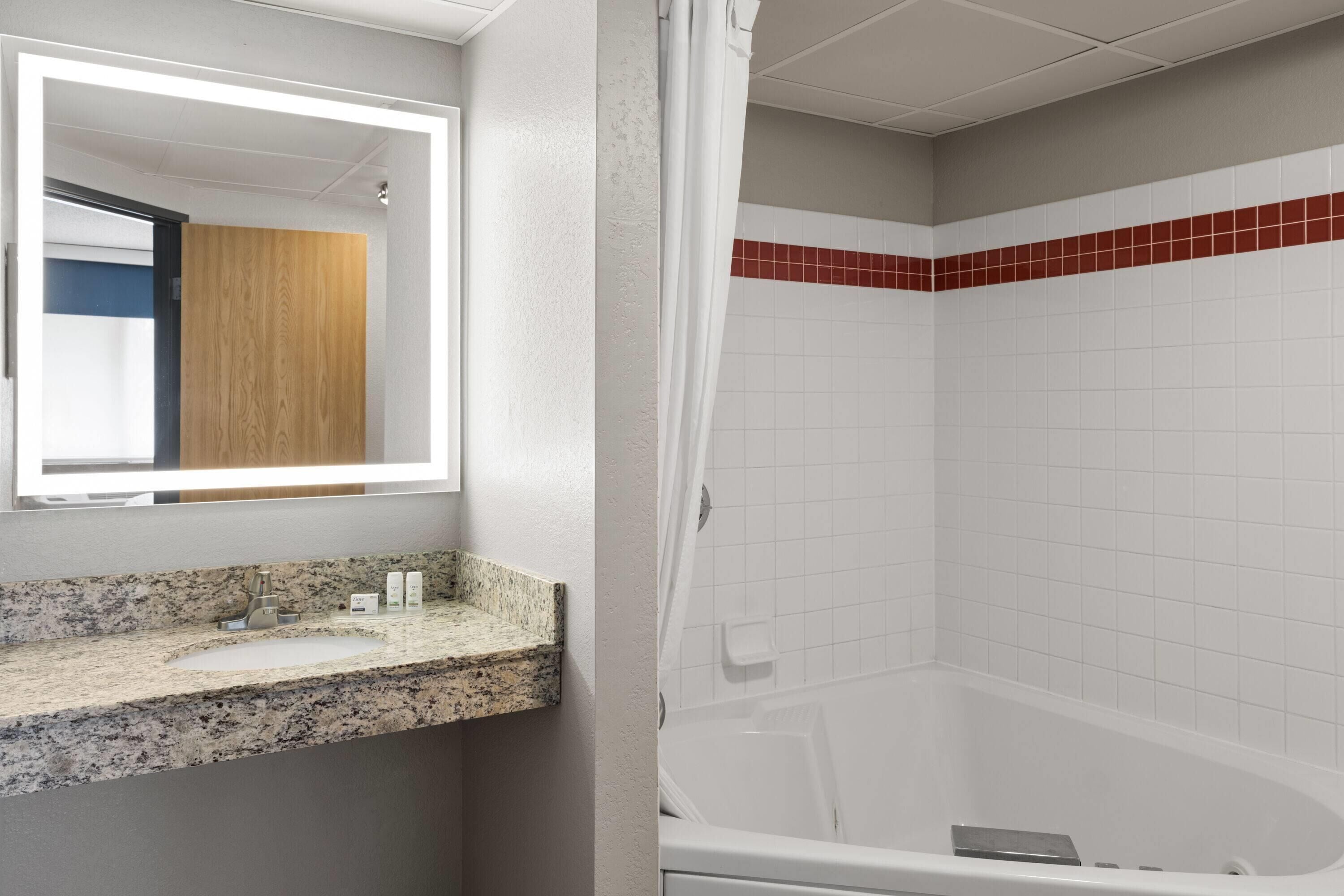 A bathroom with a granite countertop sink, a mirror, and a white bathtub with red-tiled walls.