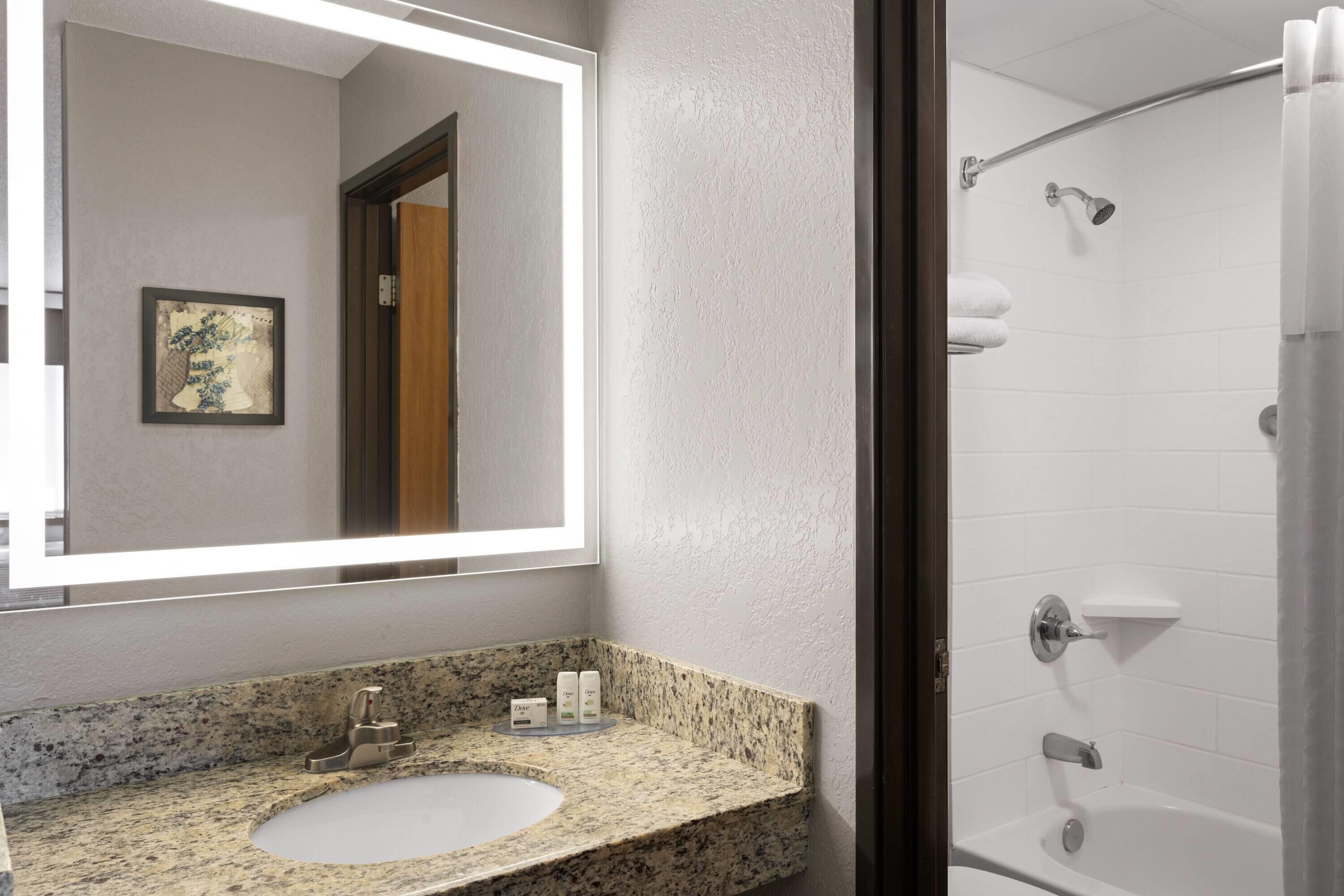 Un baño con lavamanos de encimera de granito, un espejo grande y una zona de ducha.