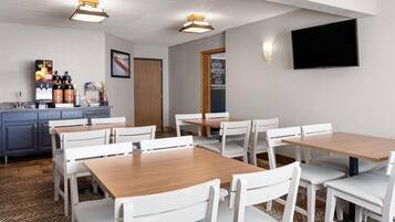 A hotel room with a dining area featuring white chairs and tables, a kitchen area with a coffee station, and a flat-screen TV mounted on the wall.