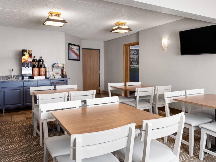 A hotel room with a dining area featuring white chairs and tables, a kitchen area with a coffee station, and a flat-screen TV mounted on the wall.