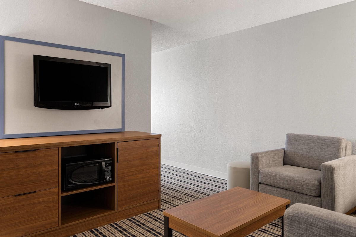 A modern hotel room with a flat-screen TV mounted on a wall, a wooden cabinet with a microwave, a grey armchair, and a wooden coffee table.