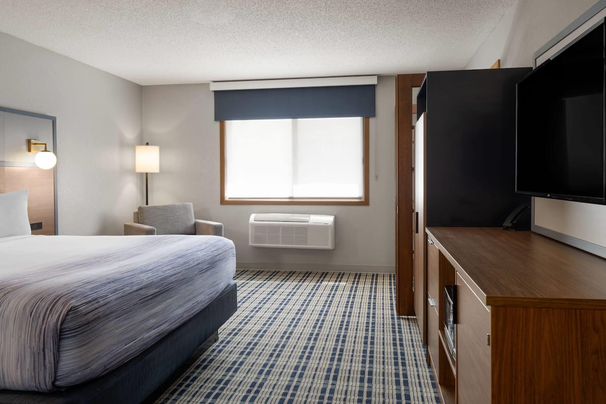 A hotel room with a bed, a desk, a television, a window with blinds, a lamp, and an air conditioning unit.