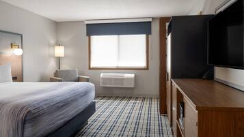Habitación de hotel con cama, escritorio, televisor, ventana con persianas, lámpara y aire acondicionado.