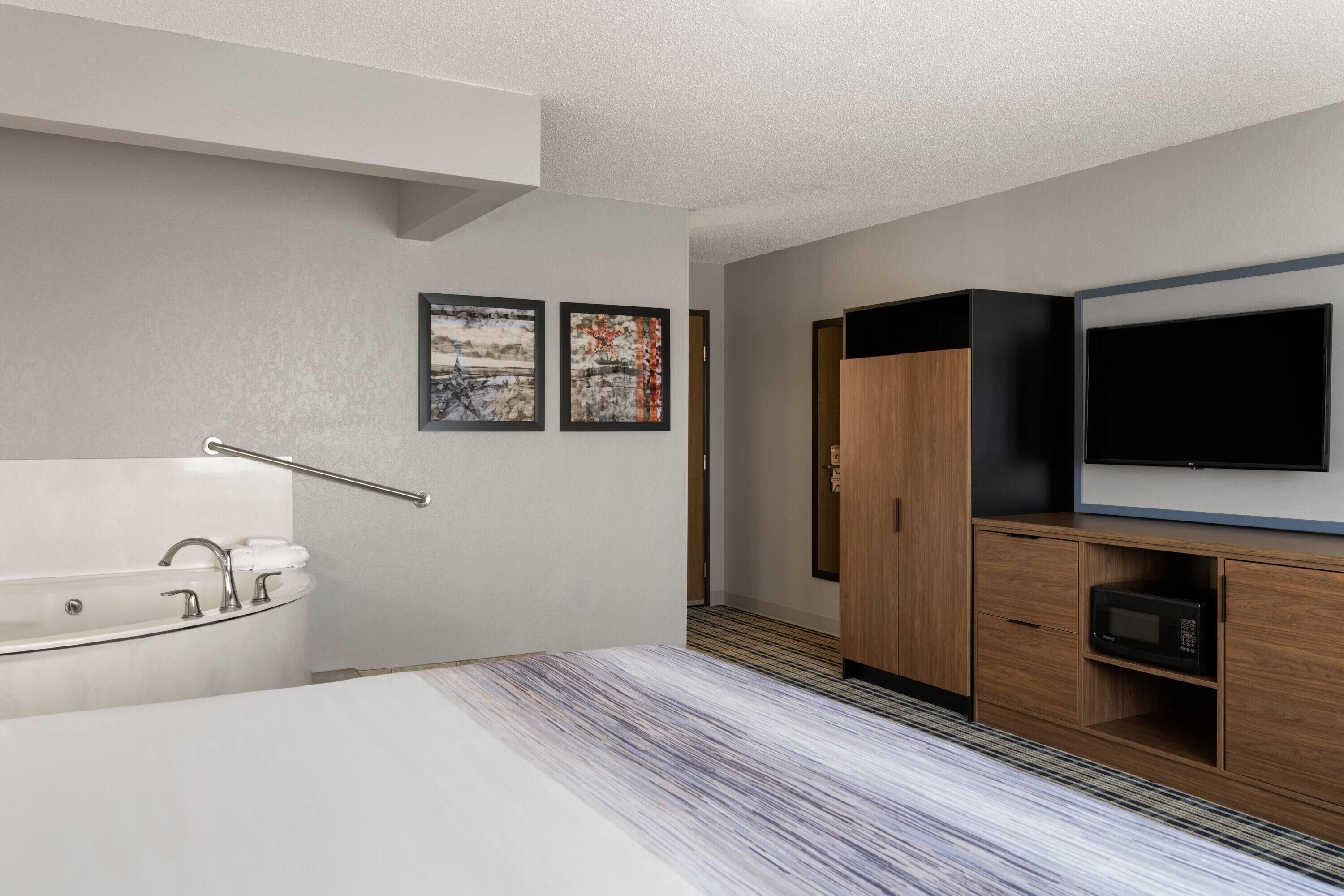 A modern hotel room with a large bed, a built-in TV, a wooden wardrobe, and a bathtub with a towel rail.