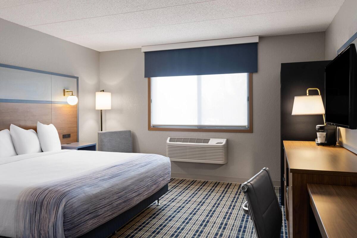 A hotel room with a large bed, a desk with a lamp, a TV, and a window with blinds.