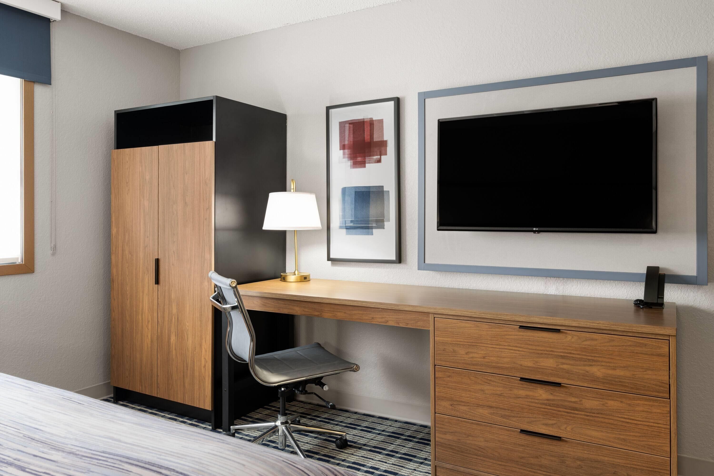 Ett hotellrum med ett skrivbord i trä, en stol, en säng, en tv och en inramad tavla på väggen.