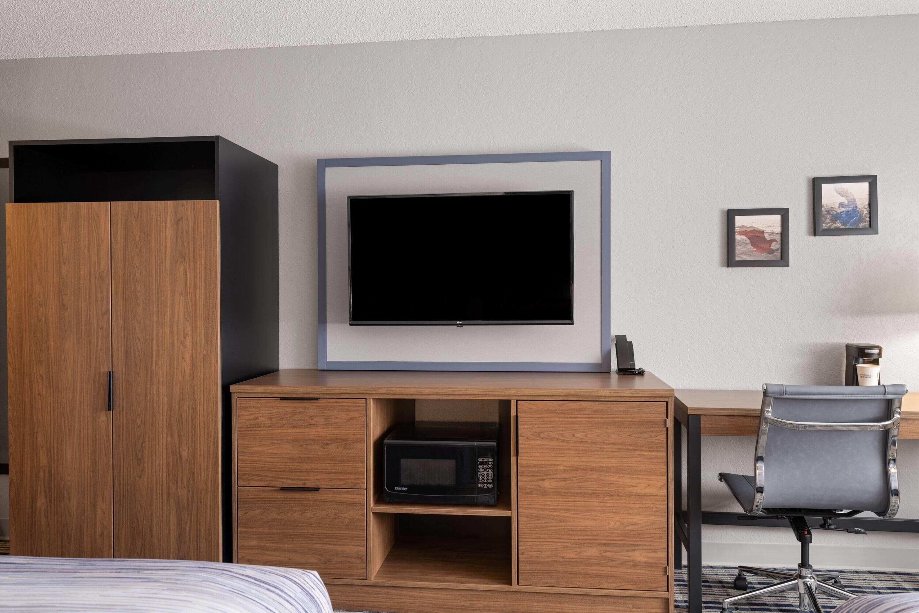A hotel room with a wooden wardrobe, a flat-screen TV mounted on a wall, a desk with a chair, and a microwave.