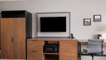 A hotel room with a wooden wardrobe, a flat-screen TV mounted on a wall, a desk with a chair, and a microwave.
