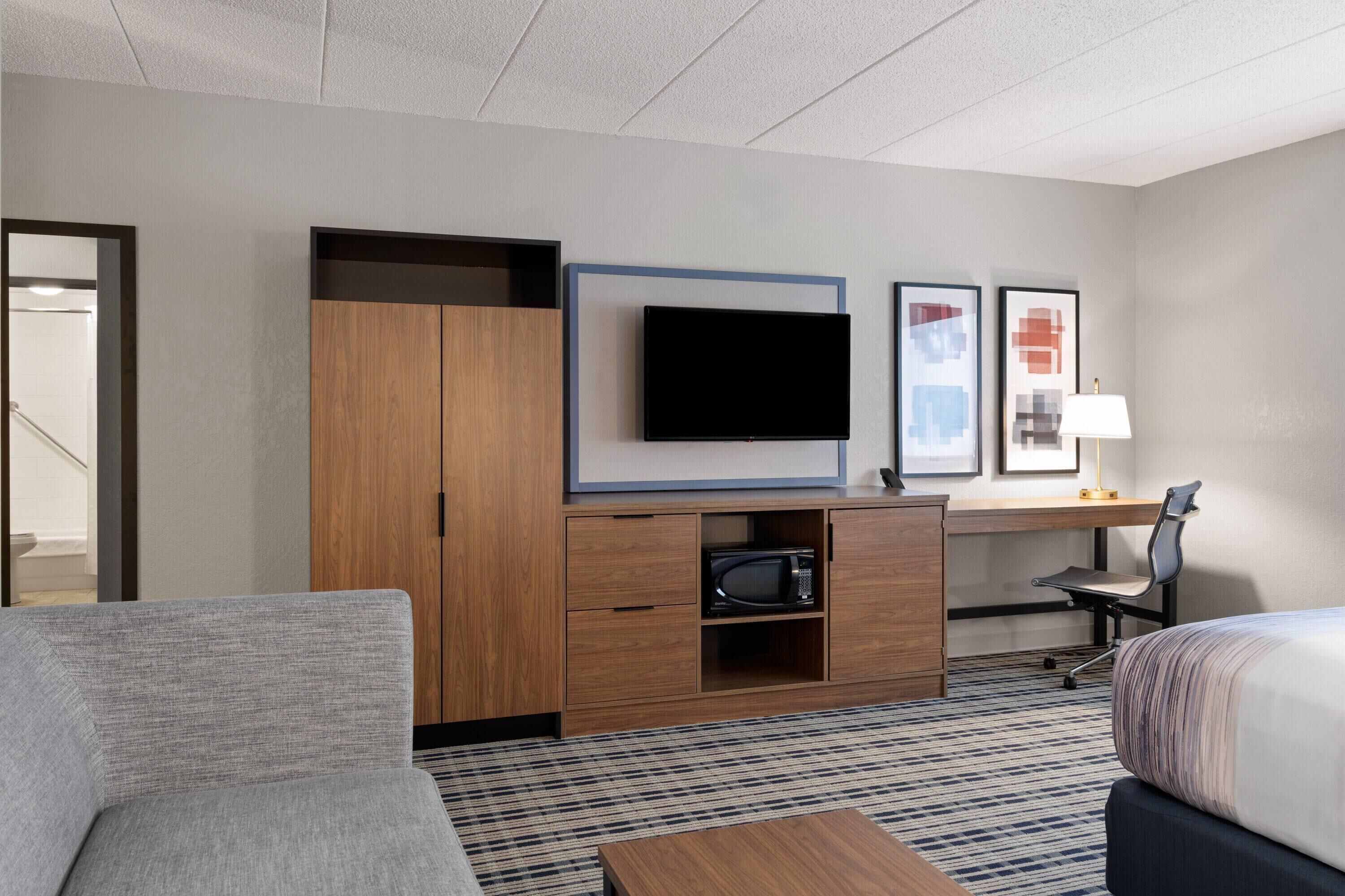 A hotel room with a sofa, a bed, a desk with a flat-screen TV, a microwave, and framed pictures on the wall.