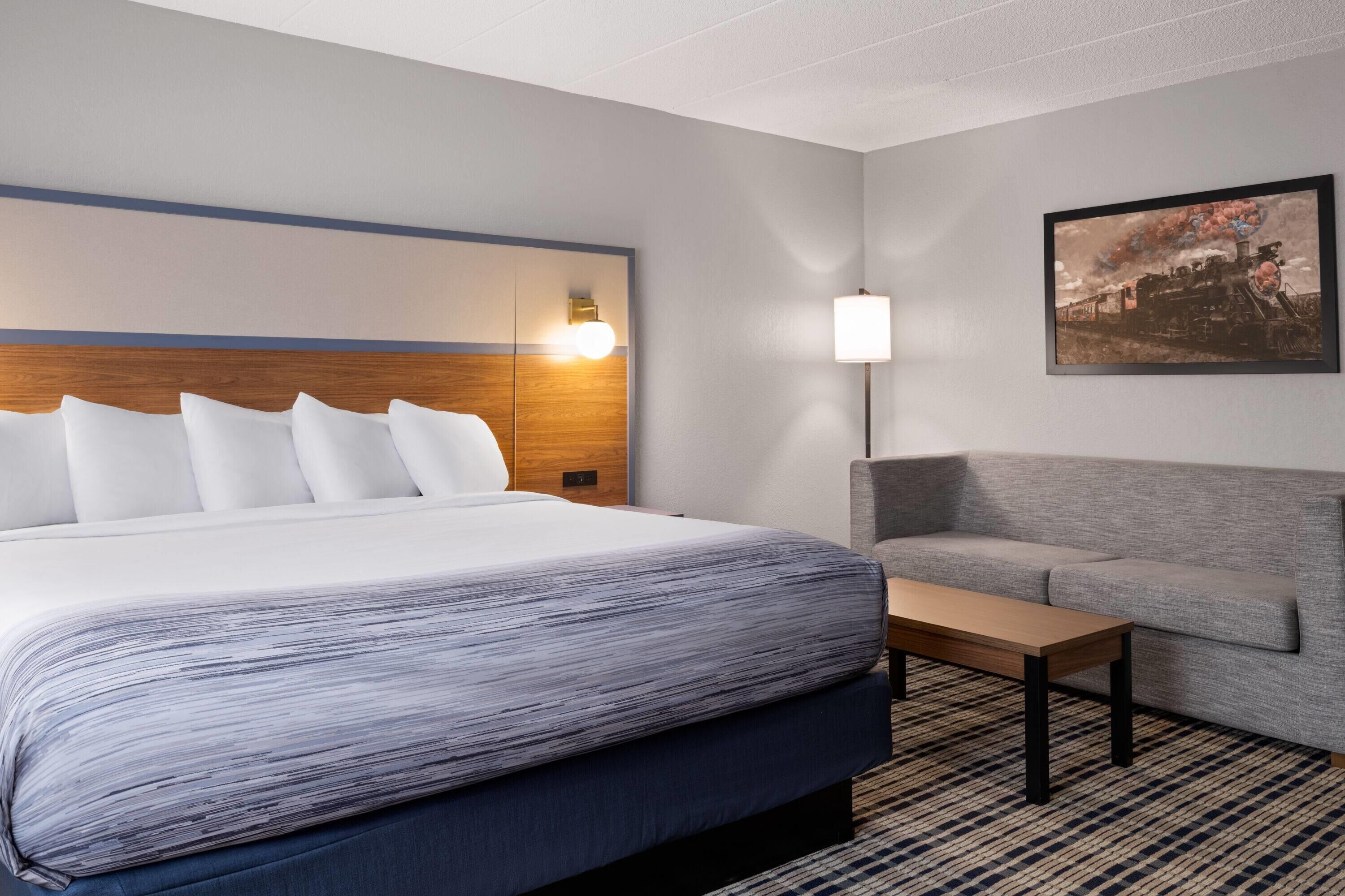 A hotel room with a large bed, a gray sofa, a wooden nightstand, and a painting on the wall.