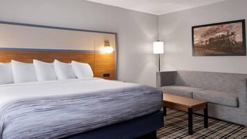 A hotel room with a large bed, a gray sofa, a wooden nightstand, and a painting on the wall.