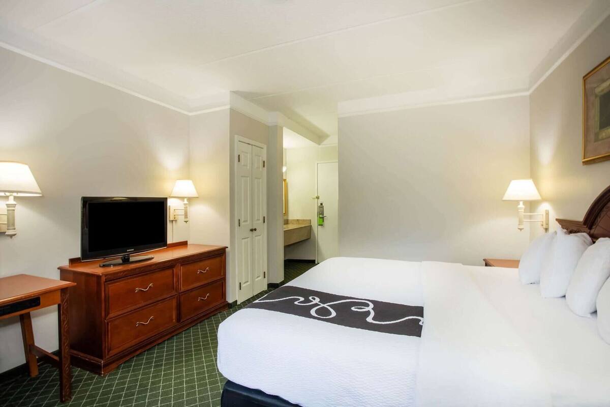 A hotel room with a bed, a TV on a wooden dresser, a bathroom visible through an open door, and two wall-mounted lamps.