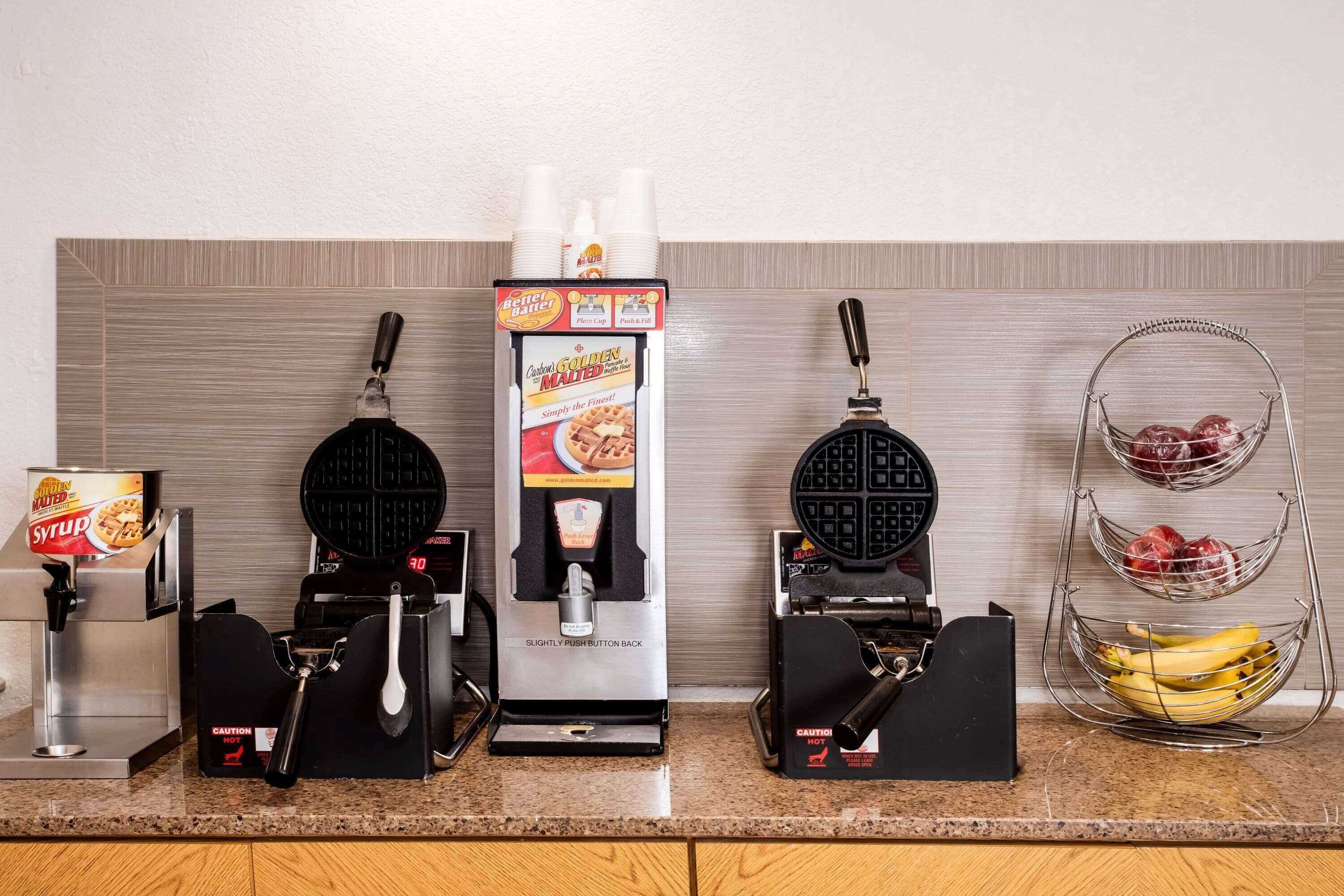 Uma bancada de cozinha com uma máquina de waffles, uma cafeteira e uma cesta de frutas.