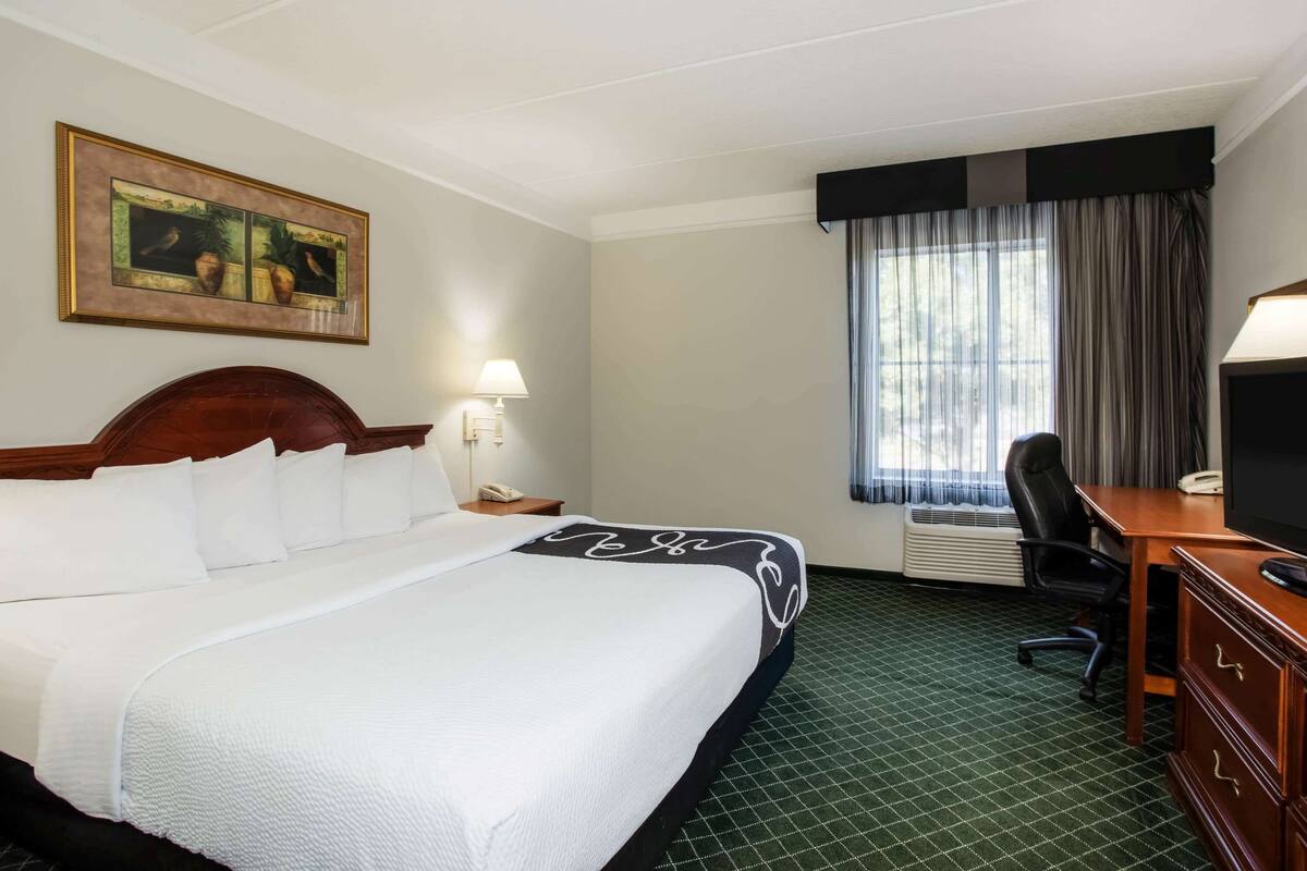 A hotel room with a large bed, a desk, a chair, a TV, a lamp, and a window with curtains.