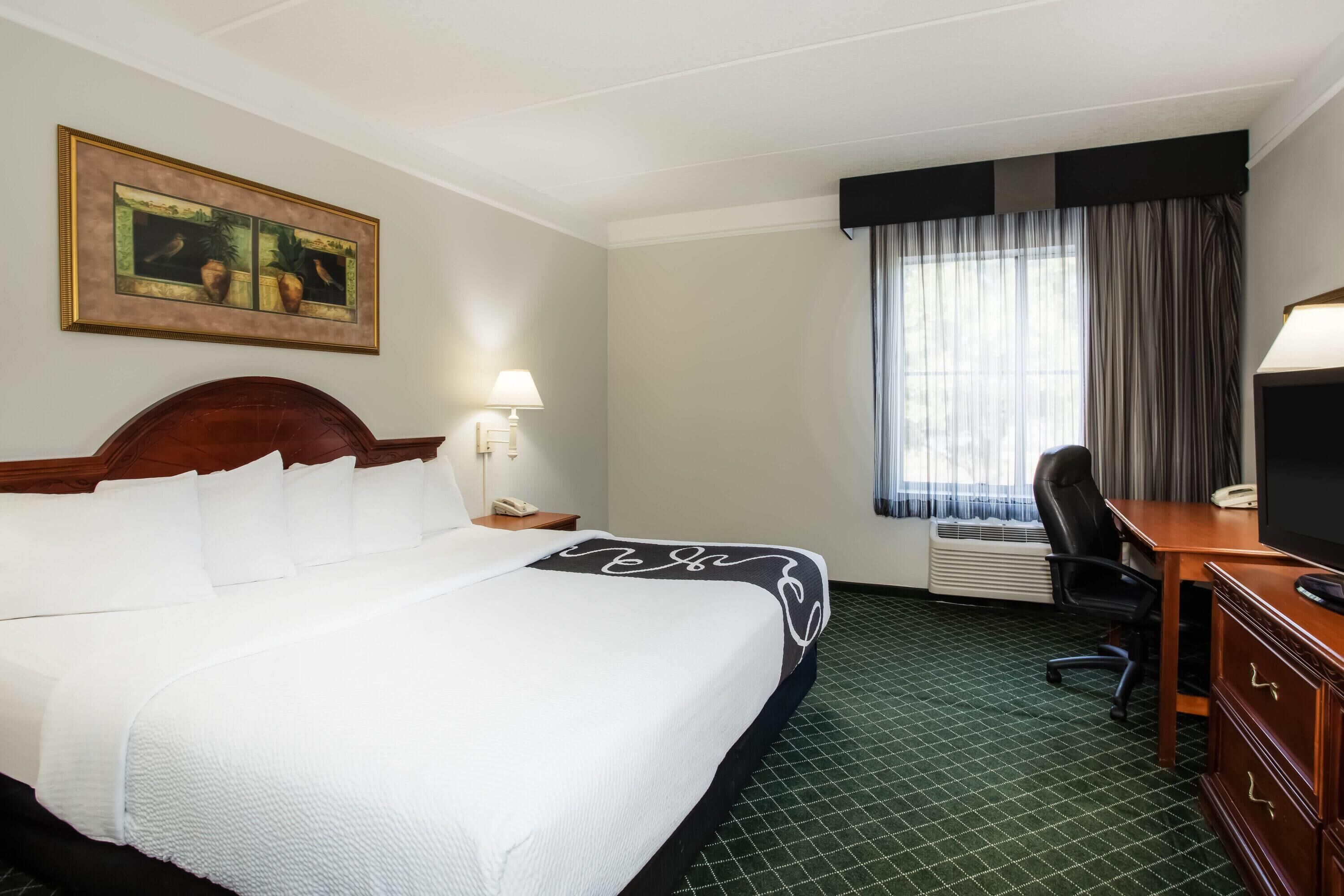 A hotel room with a large bed, a desk, a chair, a telephone, a lamp, and a window with curtains.