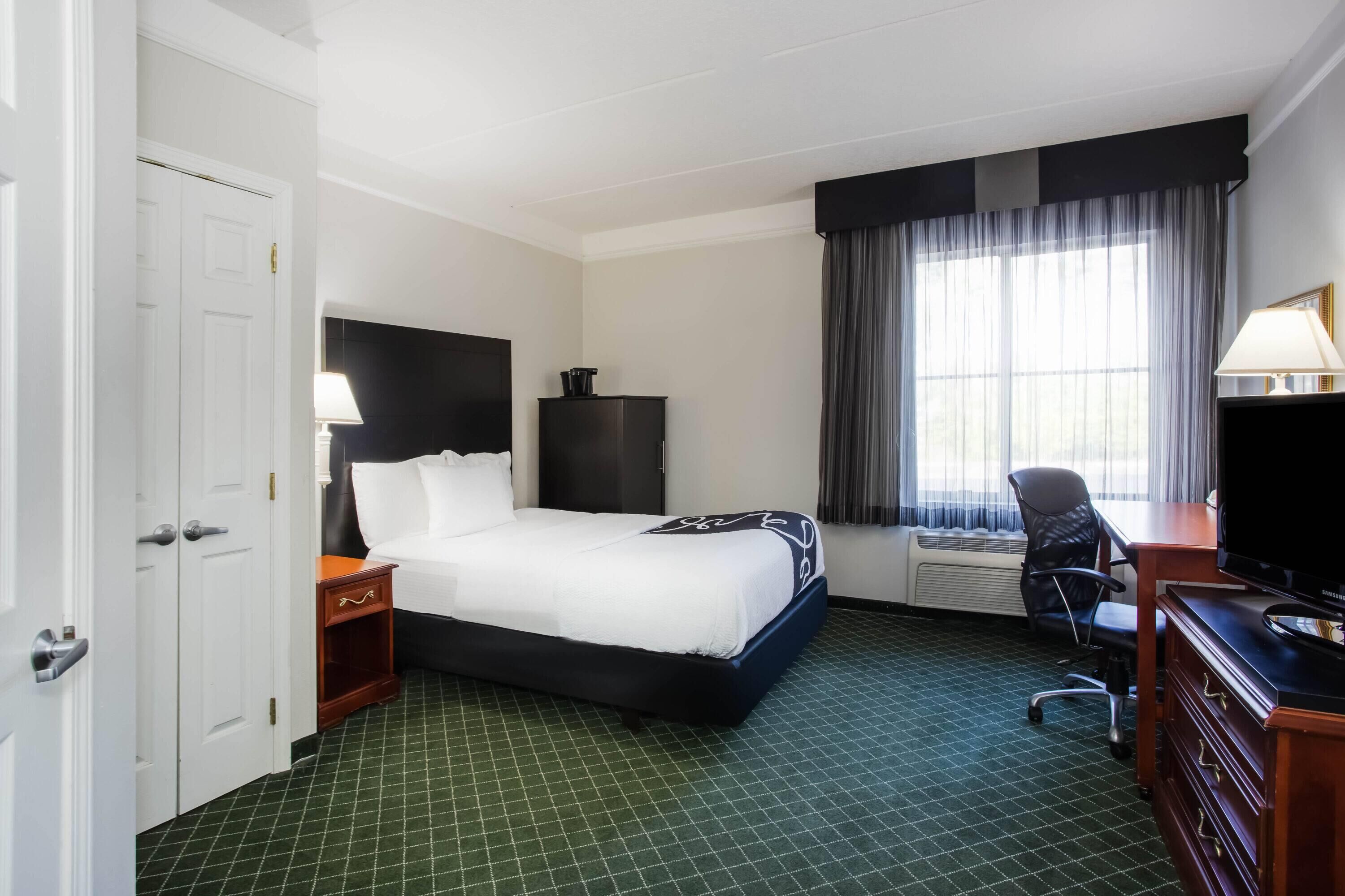 Ein Hotelzimmer mit Bett, Schreibtisch mit Computer, Stuhl, Fernseher, Fenster mit Vorhängen und einer Tür.