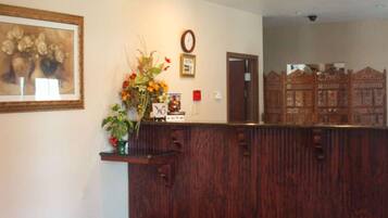 A reception area with a wooden reception desk, a framed painting of flowers, a clock, and a mirror reflecting a wooden cabinet.