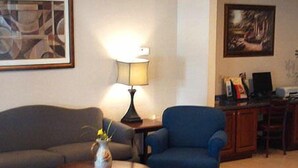A living room with a sofa, armchair, coffee table, lamp, painting, and a desk with a computer. - Western Motel Inn and Suites Hazlehurst (Hazlehurst)