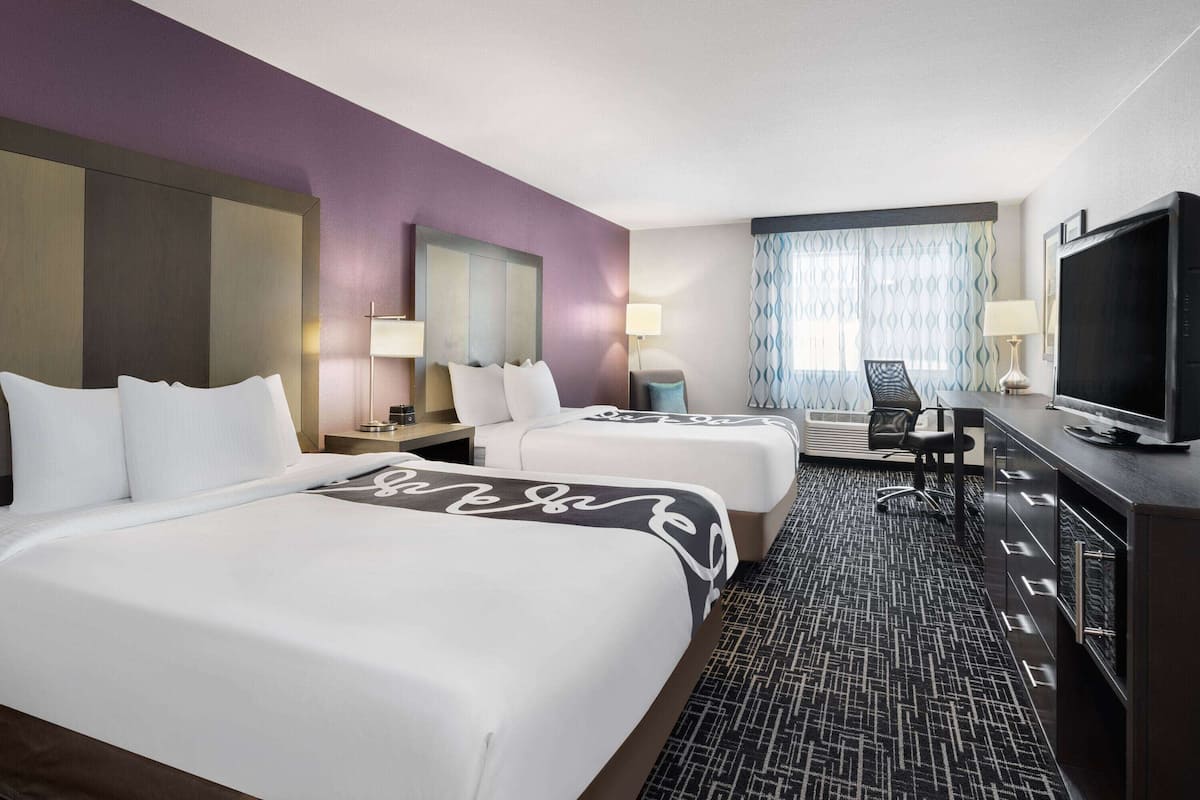 A hotel room with two beds, a desk, a television, and a window with curtains.