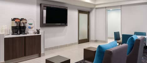 A modern hotel lobby with a reception desk, coffee machines, a flat-screen TV, and comfortable seating.