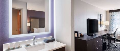 Un baño de hotel con un espejo grande, un lavamanos con dispensadores de jabón, un tocador con un televisor y un escritorio con una silla.