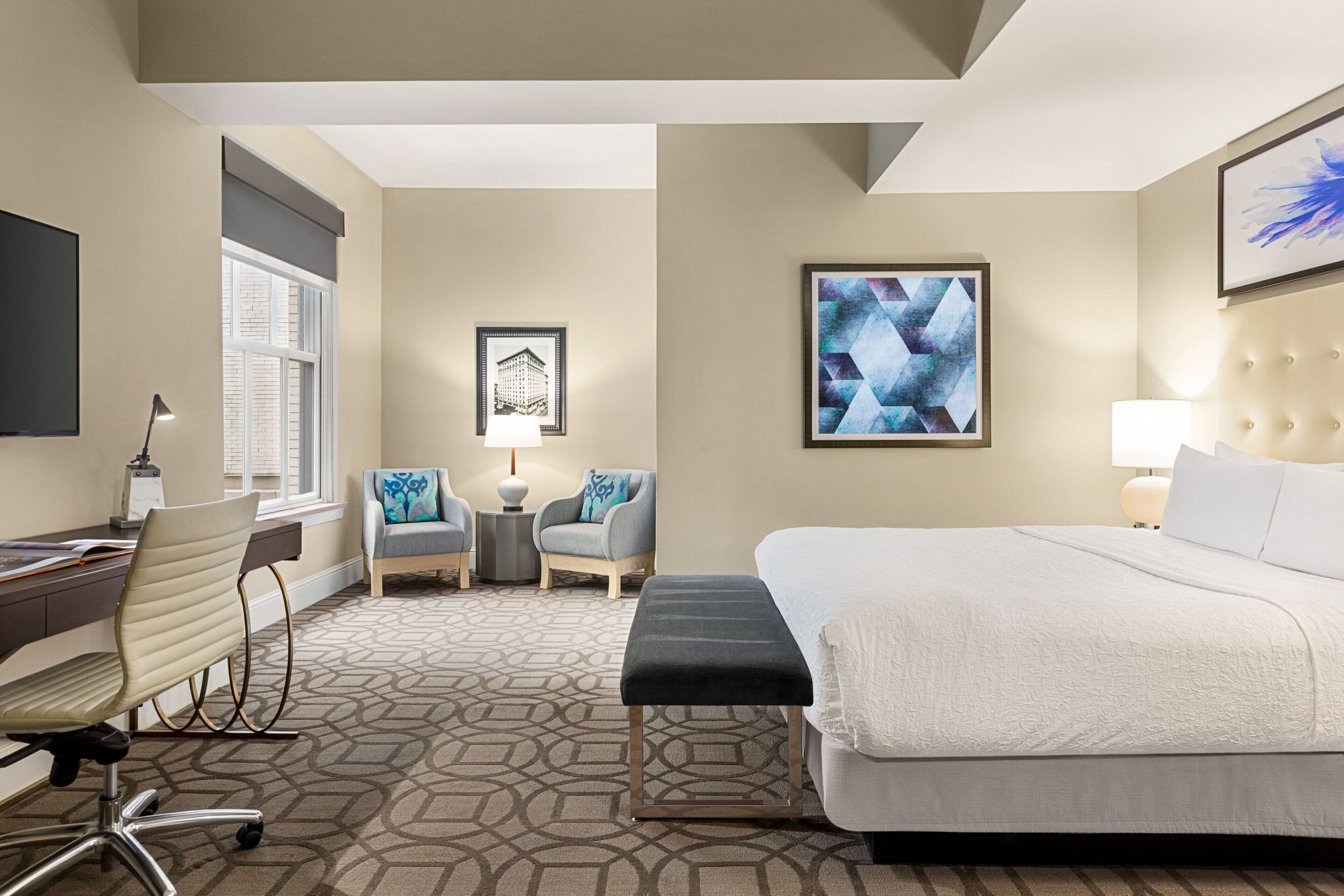 A hotel room with a bed, a desk, a chair, a small table, and two armchairs.