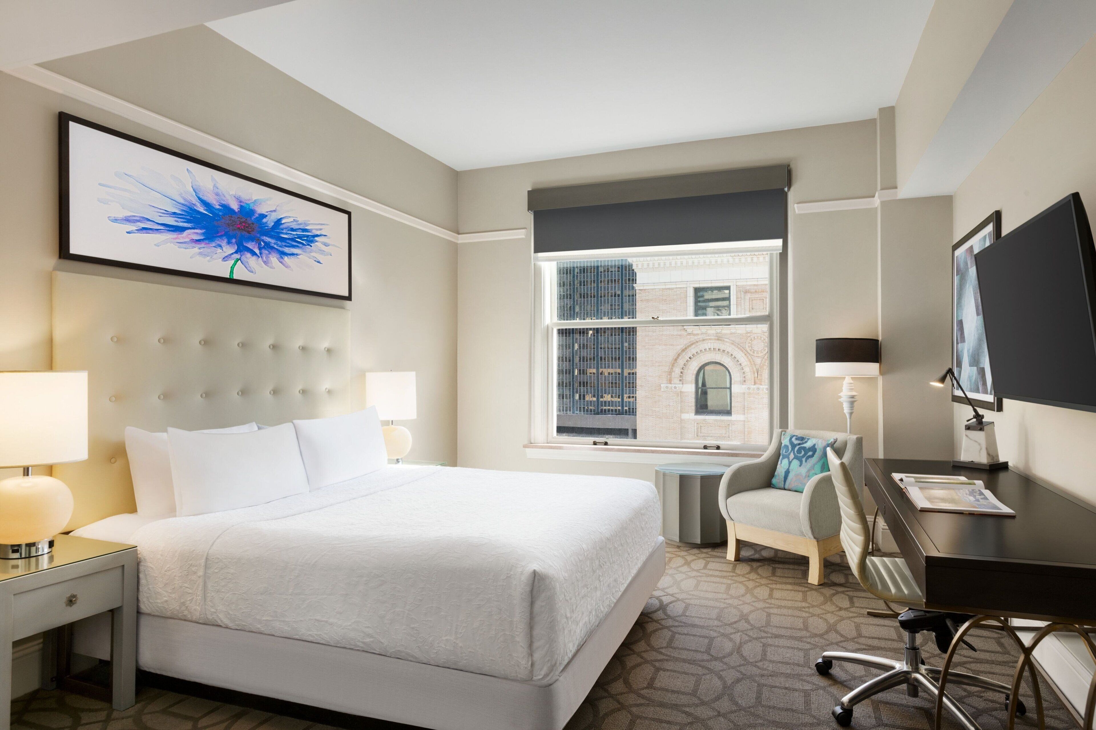 A hotel room with a large bed, a desk, a chair, a TV, and a window with a view of a building.