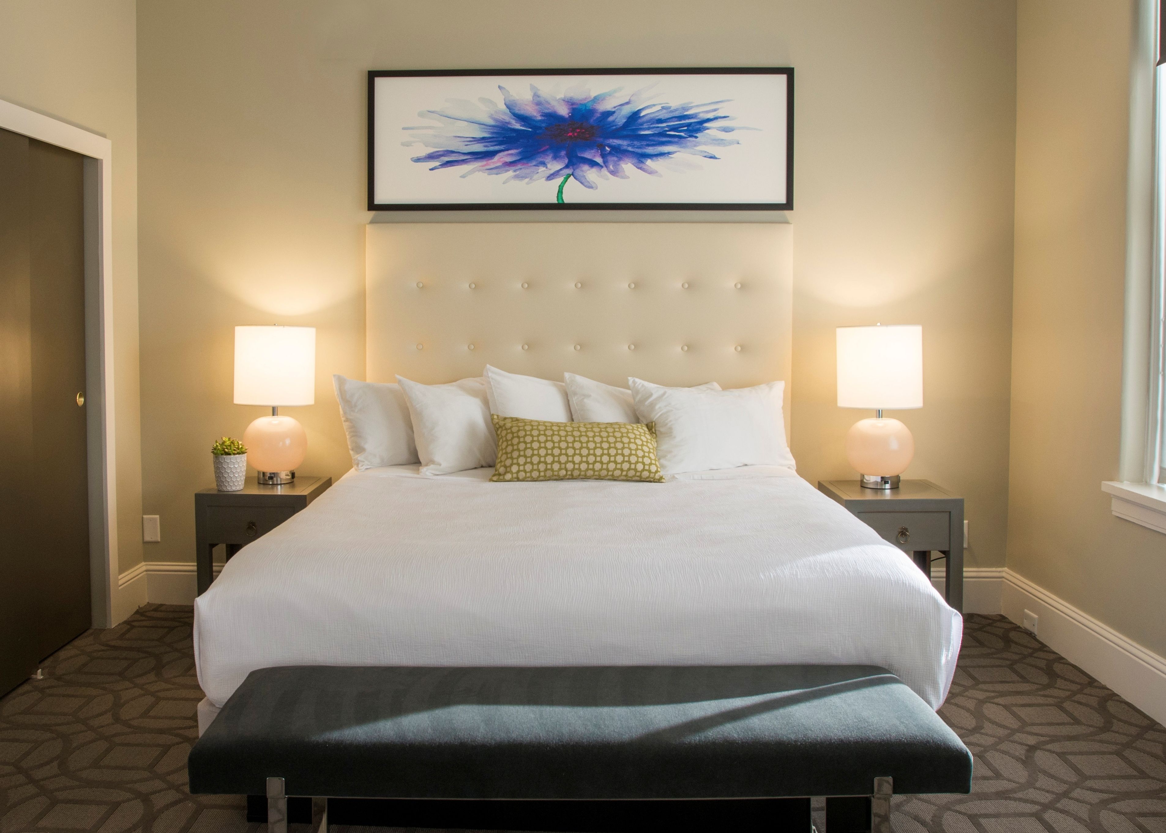 A hotel room with a large bed, two bedside tables, a bench, a framed artwork above the bed, and a window on the right.