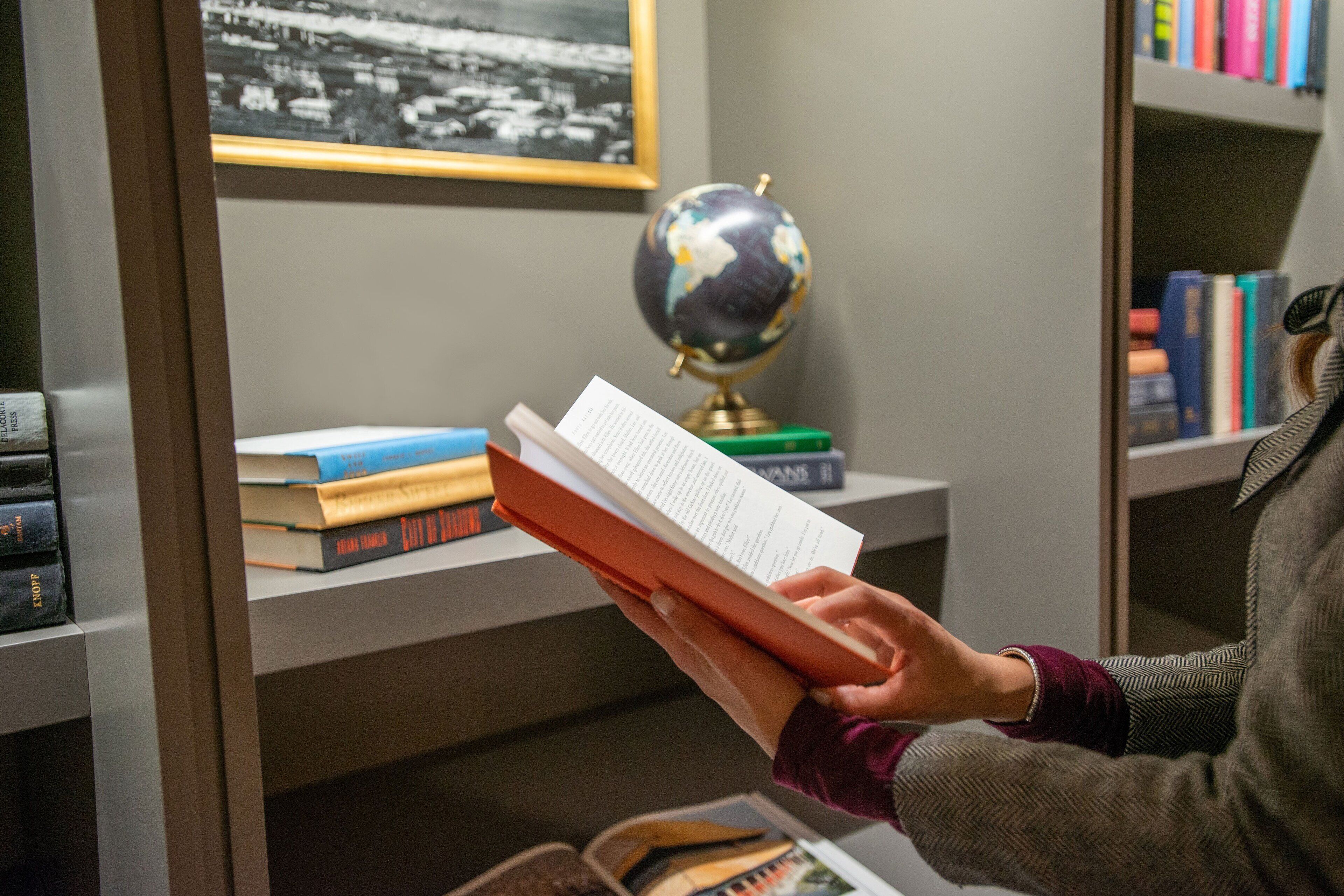 A person reading a book in a library with bookshelves, a globe, and a framed picture on the wall.
