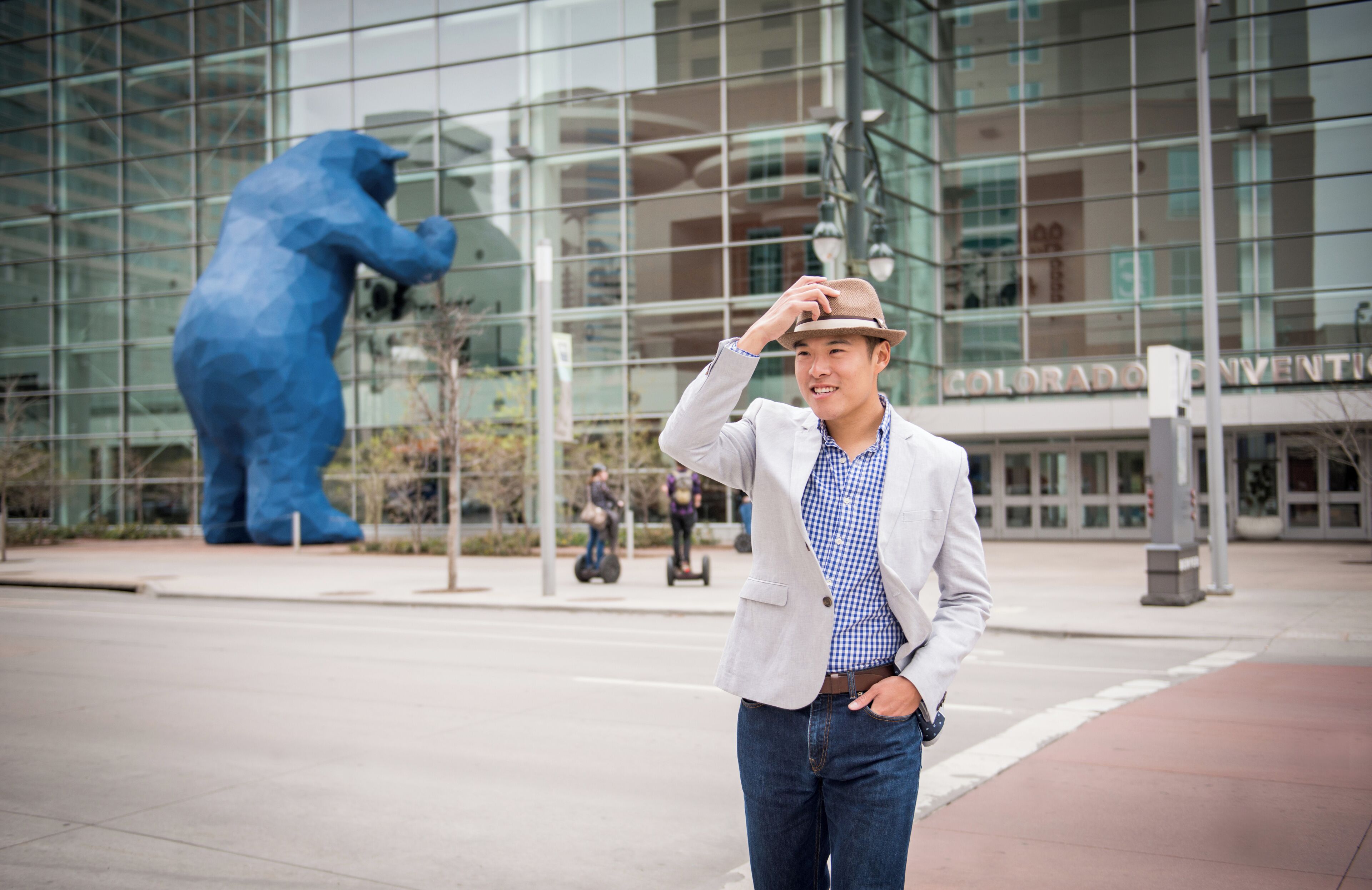 A man in a light-colored blazer and jeans standing in front of a building with a large blue bear sculpture.