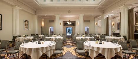 A large banquet hall with multiple round tables, high ceilings, and a central chandelier.
