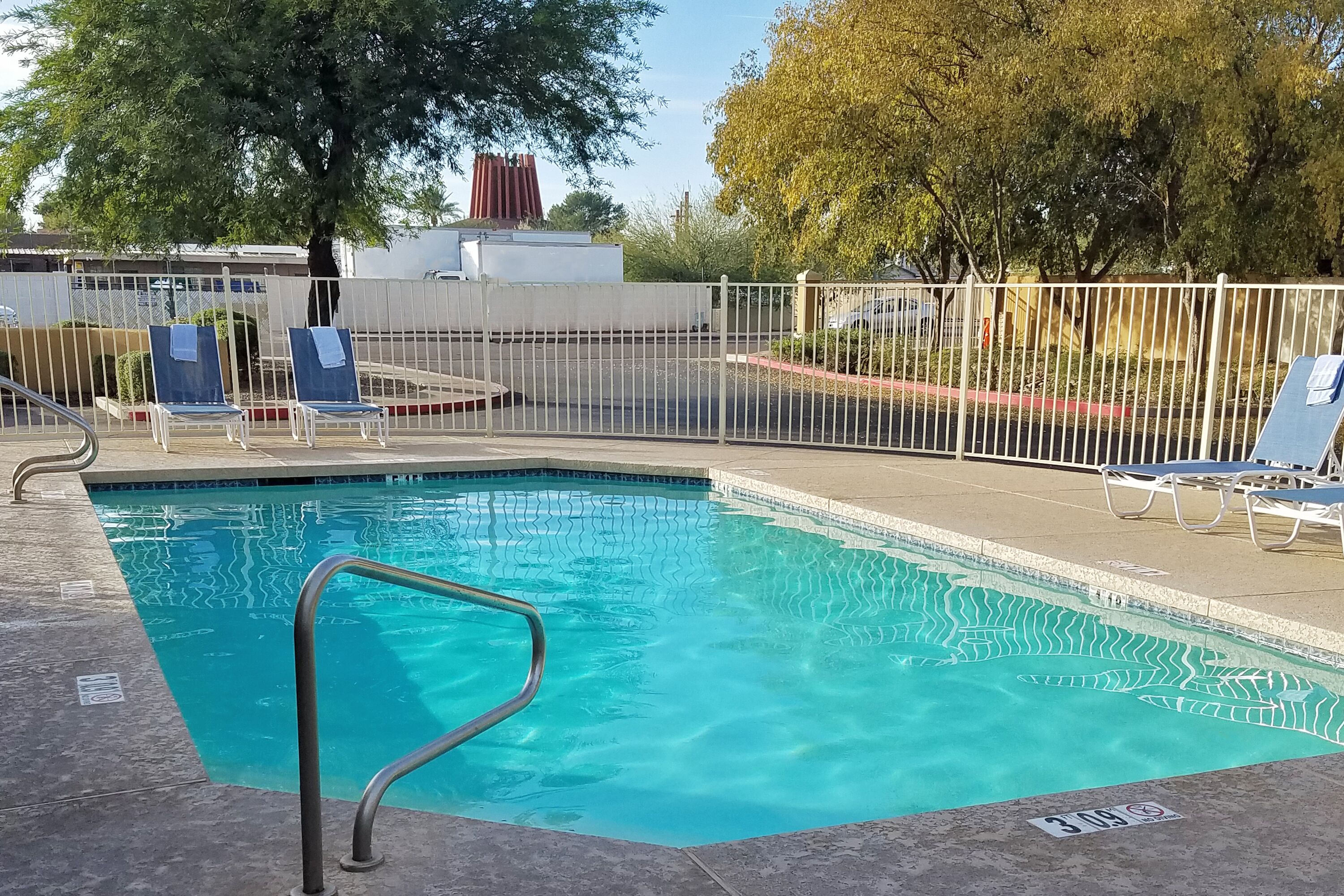 A swimming pool with a metal safety railing, surrounded by a white fence and blue lounge chairs.