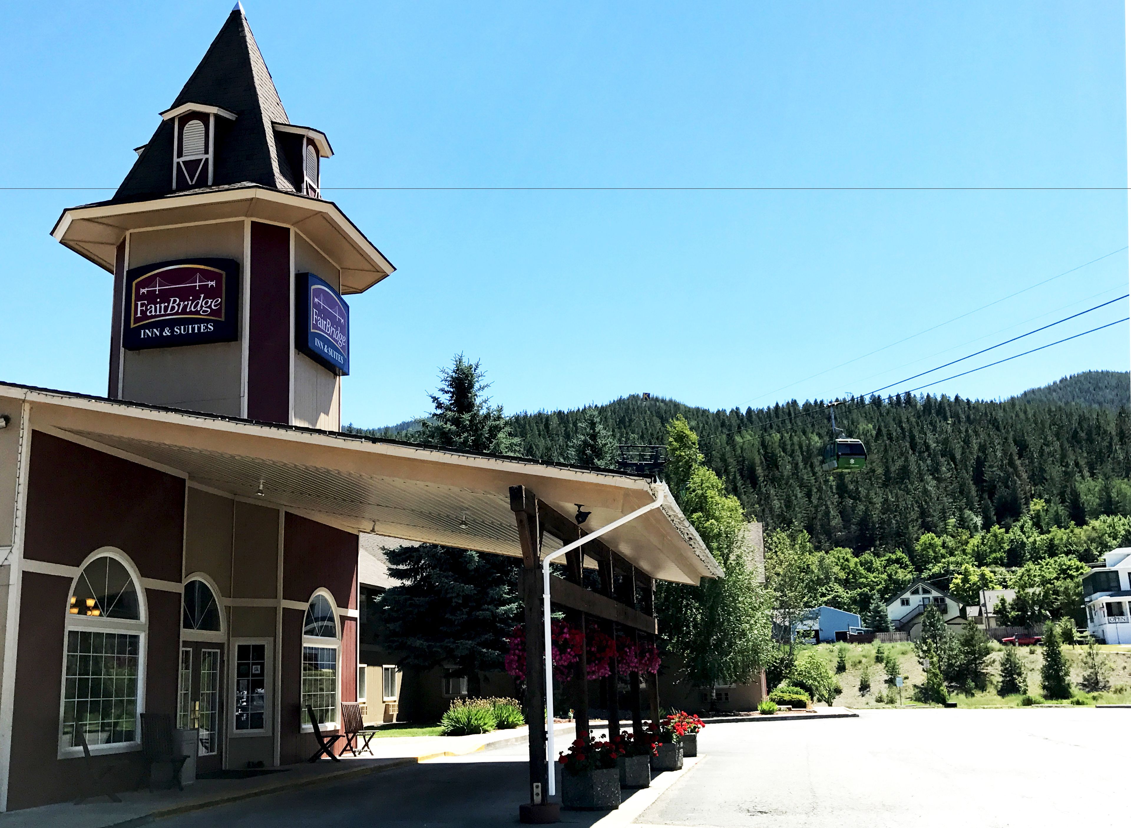 A FairBridge Inn & Suites with a prominent tower, outdoor seating, and a view of a forested mountain.