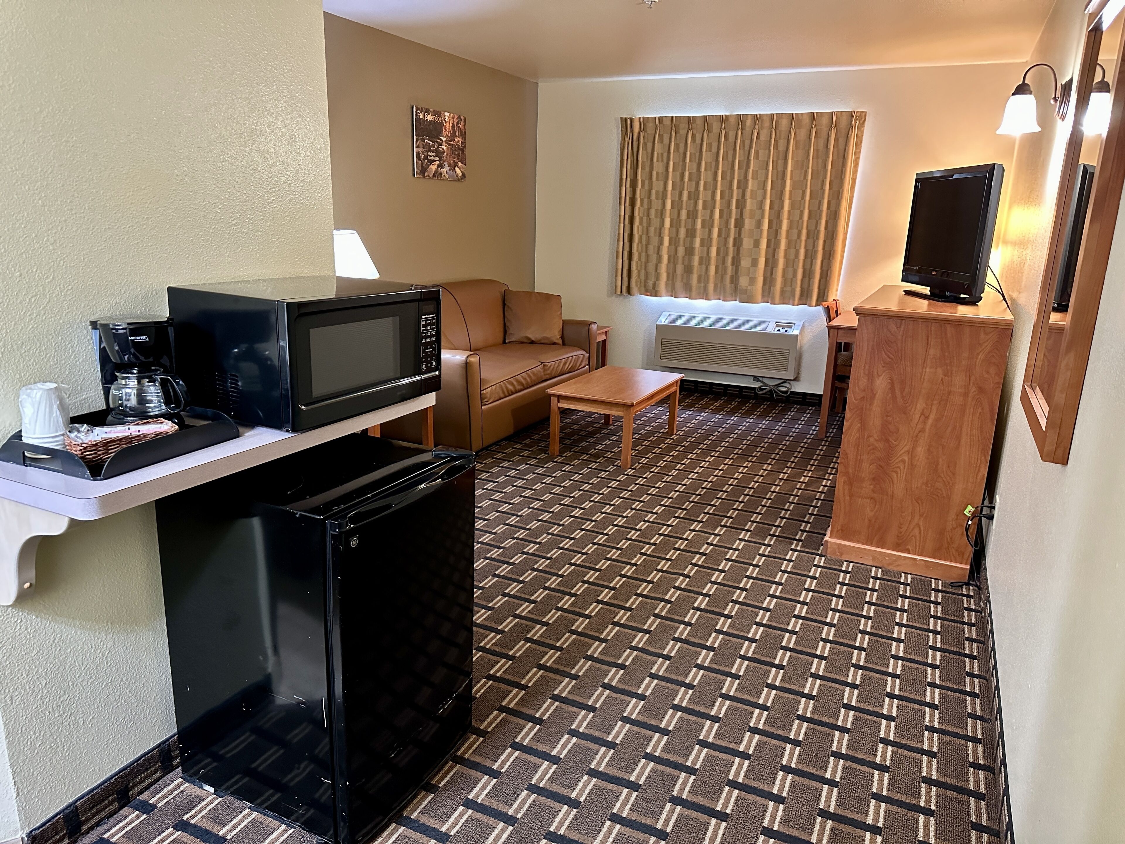 Habitación de hotel compacta con kitchenette, un sofá, una mesita de centro, un televisor y una alfombra estampada.