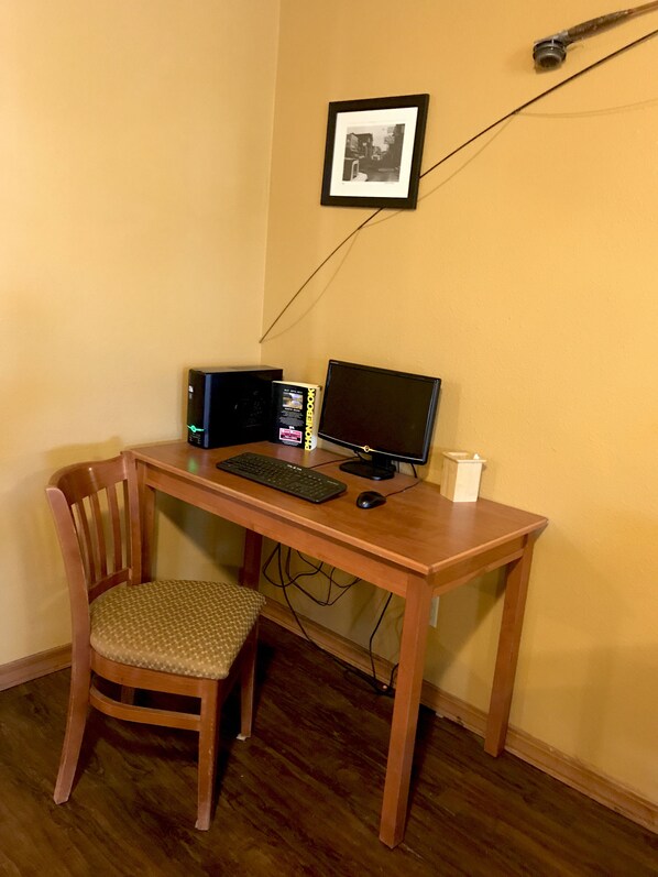 A wooden desk with a computer setup including a monitor, keyboard, and mouse. There is a chair in front of the desk and a framed picture on the wall.