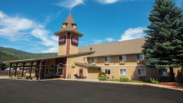 Un motel con una torre central, estacionamiento techado y montañas al fondo.