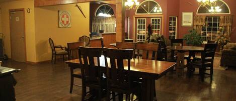 A dining area with wooden tables and chairs, a large window with curtains, and a chandelier.