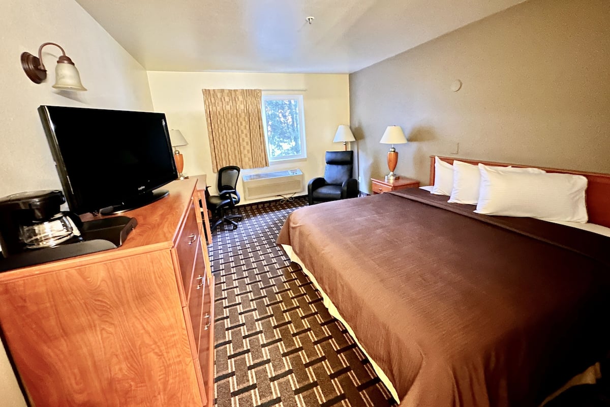 Habitación de hotel con cama, escritorio con televisor, silla, lámpara y ventana con cortinas.