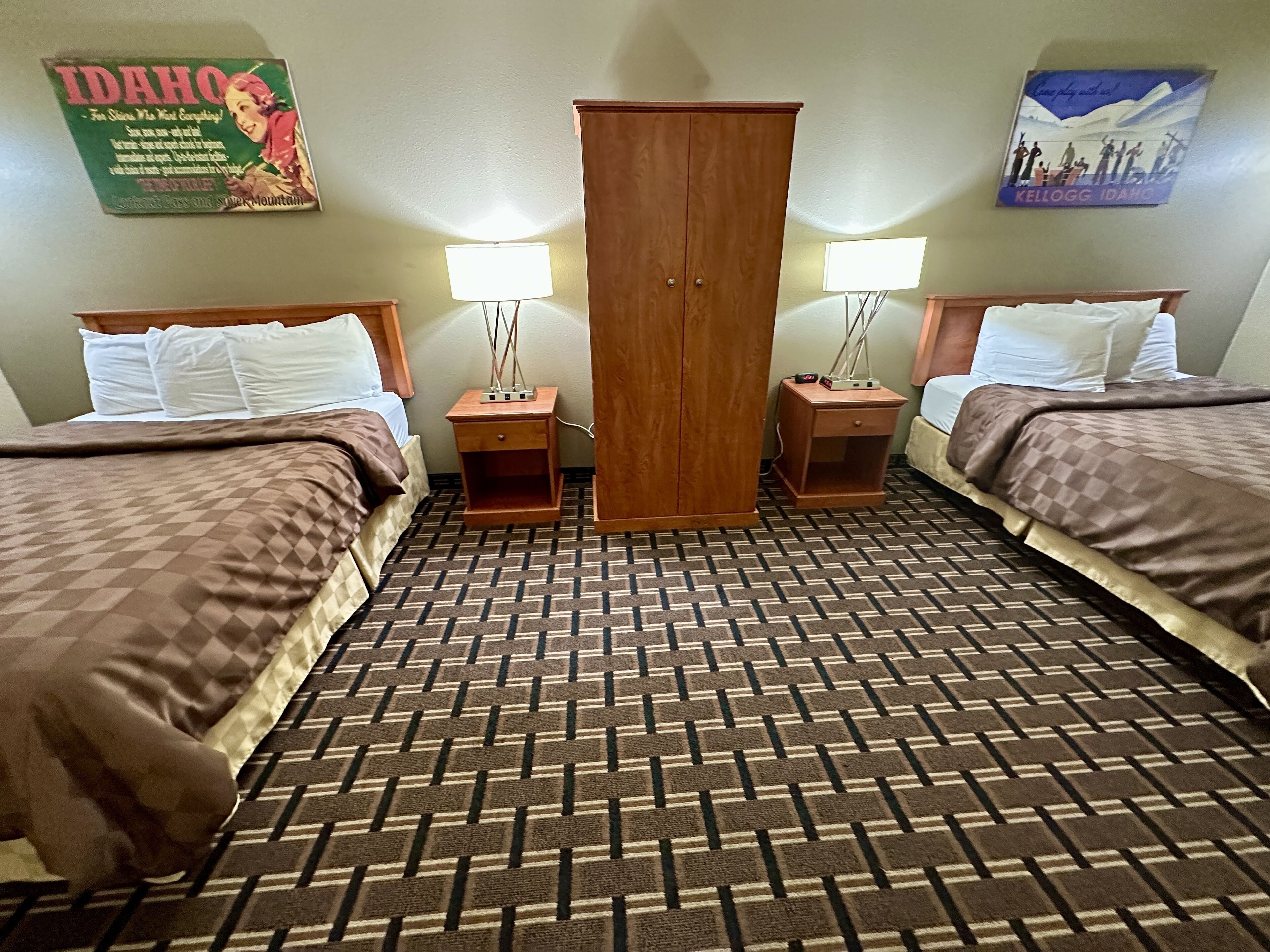 A hotel room with two beds, a wardrobe, a nightstand with a lamp, and a patterned carpet.