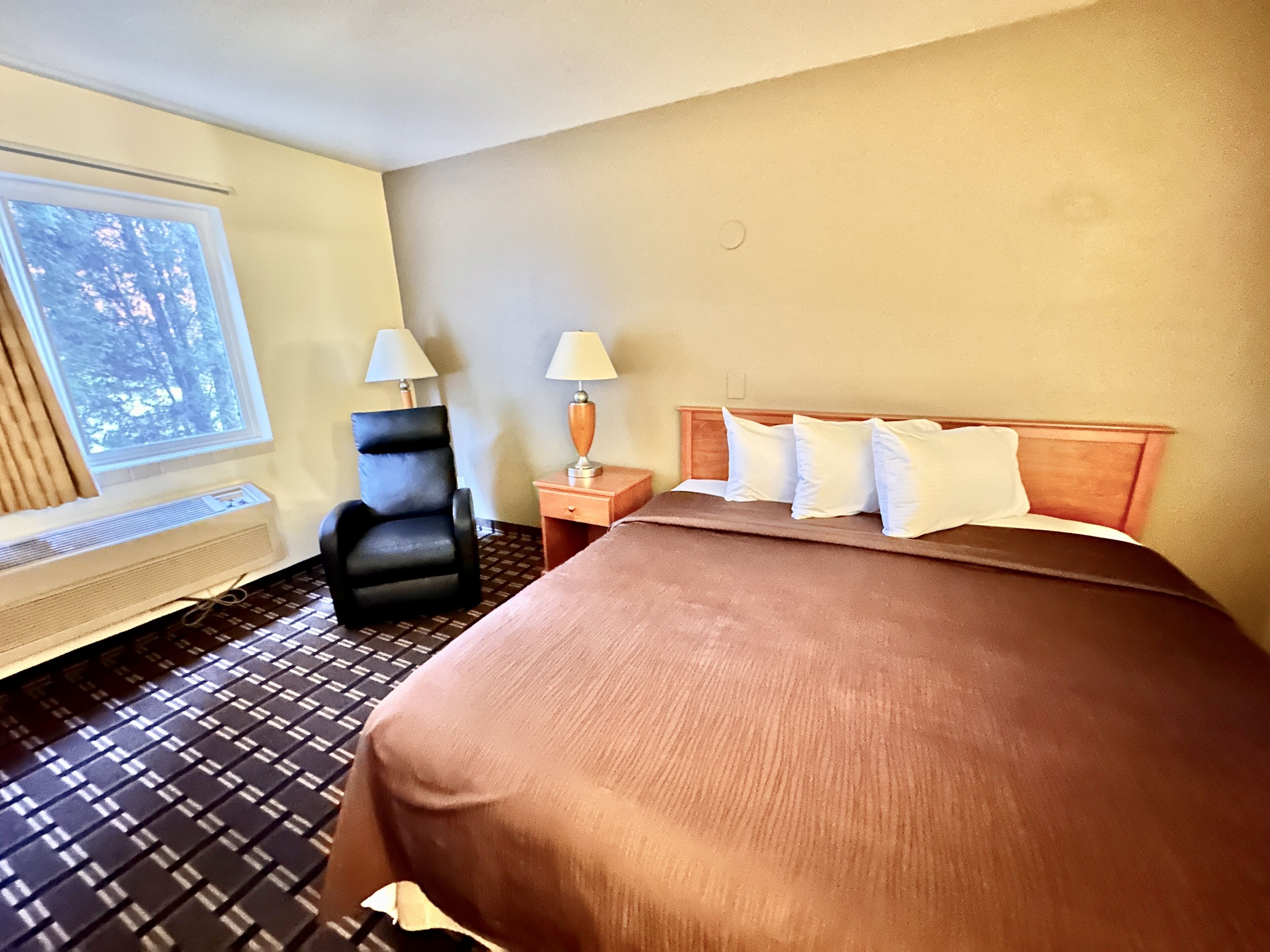 Une chambre d’hôtel avec un lit, une chaise, une table de chevet, une fenêtre et un radiateur fixé au mur.