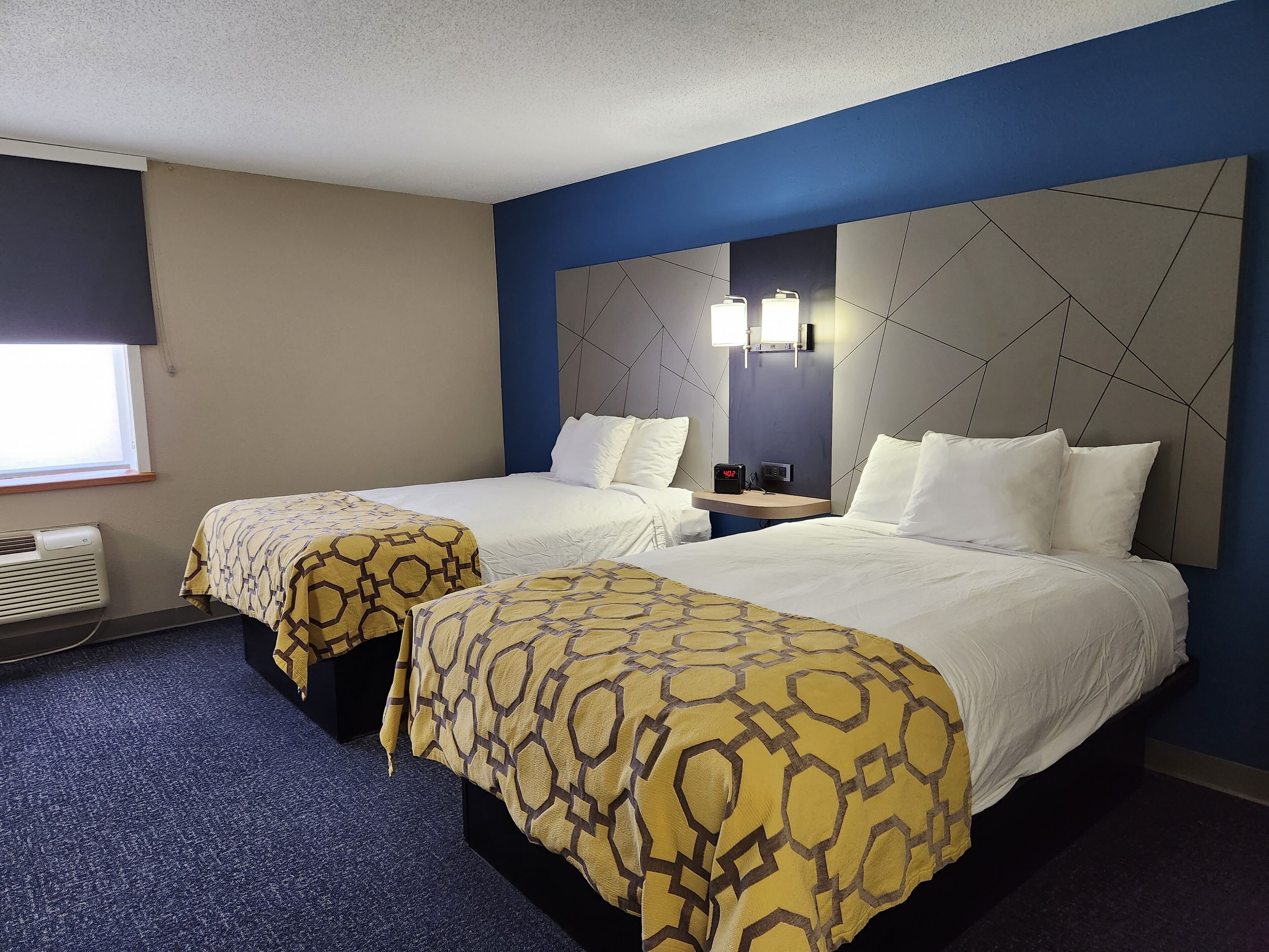 A hotel room with two beds, each with a patterned bedspread, a nightstand with a clock, and a large wall-mounted mirror.
