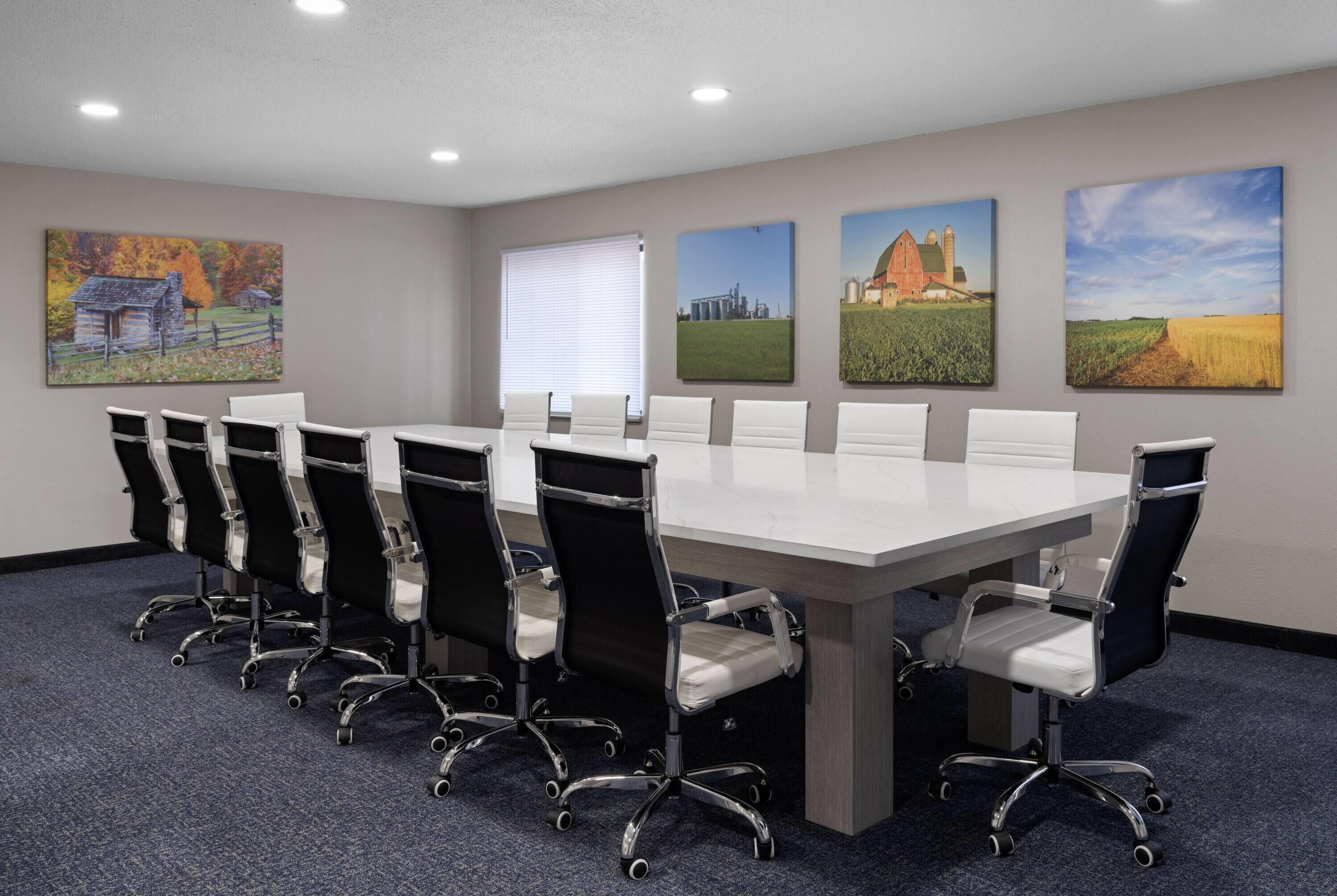 A conference room with a long table and eight chairs, featuring a cityscape, barn, and field painting on the walls.