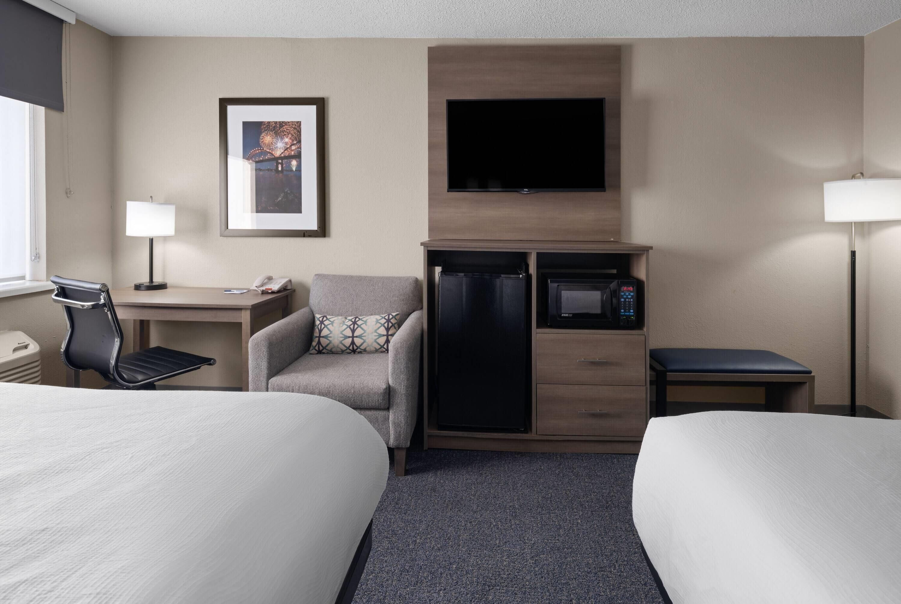 Habitación de hotel con dos camas, un escritorio con teléfono, microondas, refrigerador, una silla, un cuadro en la pared y un televisor sobre el microondas.