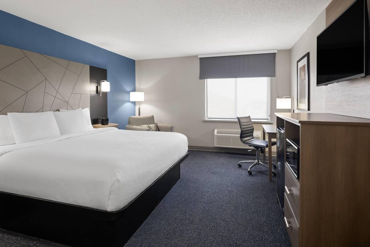A hotel room with a large bed, a desk, a chair, a TV, and a window with blinds.