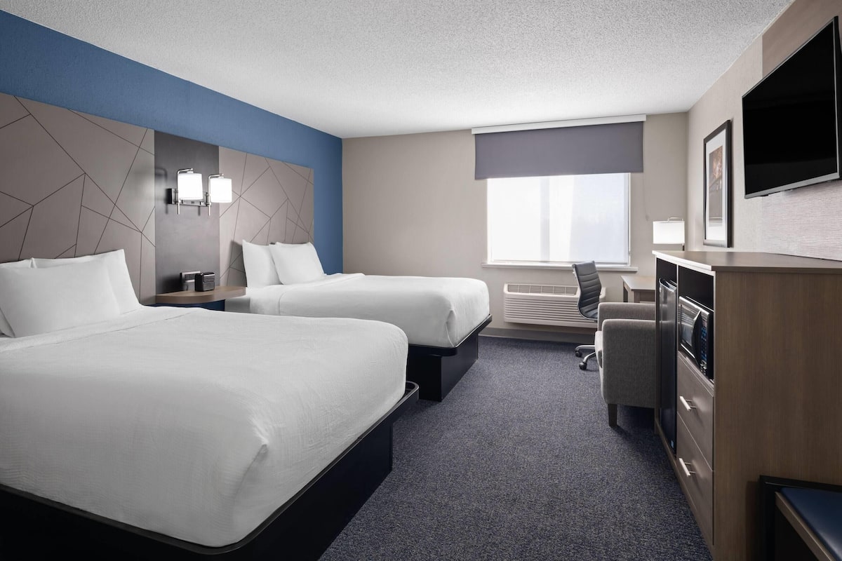 A hotel room with two beds, a desk, a chair, a flat-screen TV, and a window with a view.