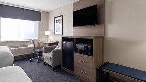 A hotel room with a bed, a desk with a chair, a microwave, a TV, and a bench.