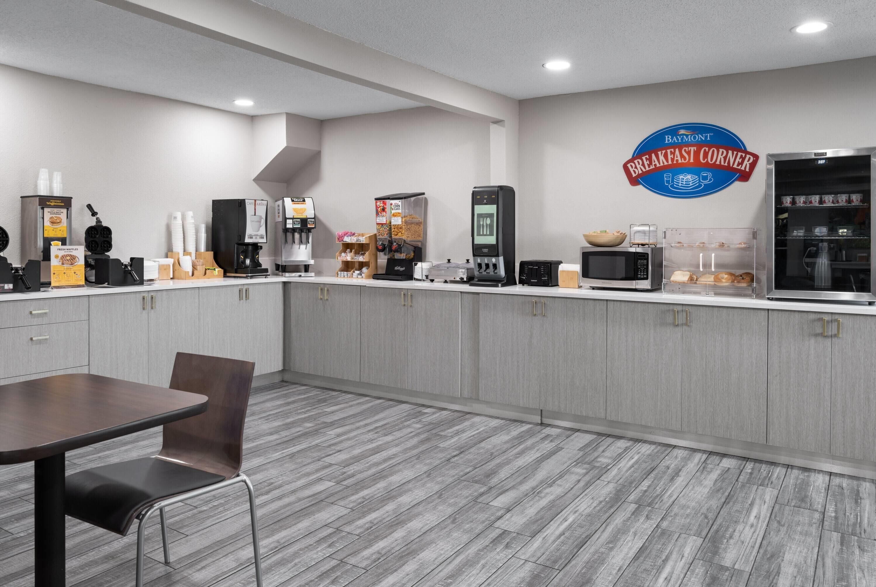 Un espace moderne dédié au petit-déjeuner, avec un comptoir, des machines à café et un panneau indiquant « Coin petit-déjeuner Baymont ».
