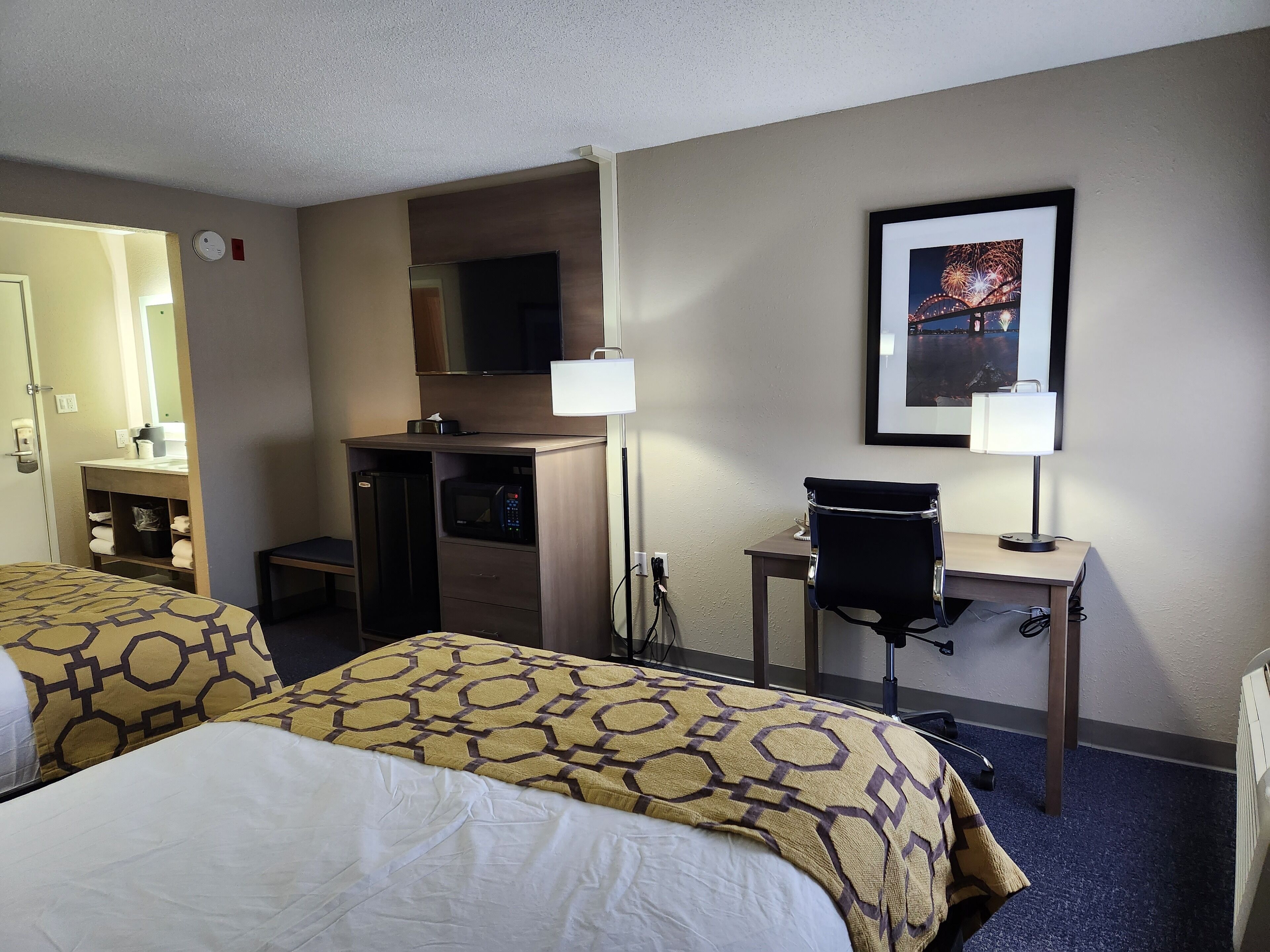 A hotel room with two beds, a desk, a chair, a microwave, a television, and a framed picture on the wall.