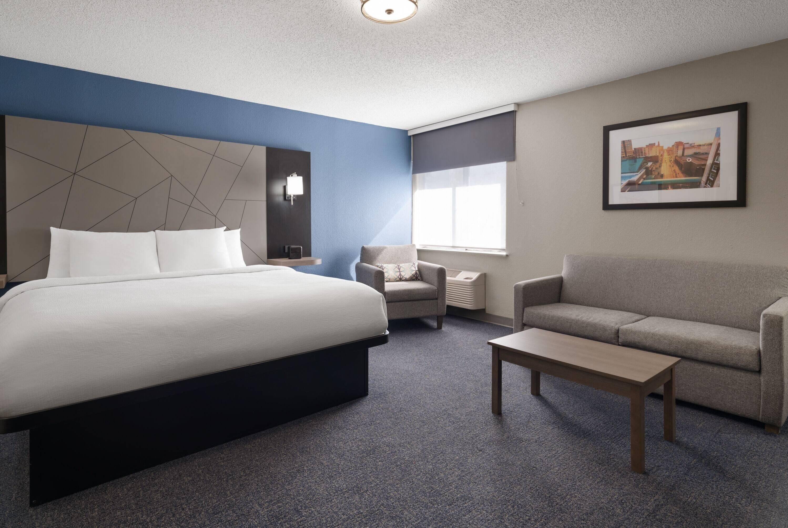 Habitación de hotel con una cama grande, un sofá, una silla, una mesita y un cuadro en la pared.